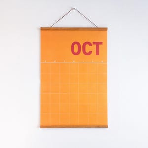 May include: An orange wall calendar for October, featuring a grid layout and the word "OCT" in red. The calendar is suspended by a wooden bar and a string, ideal for home or office.