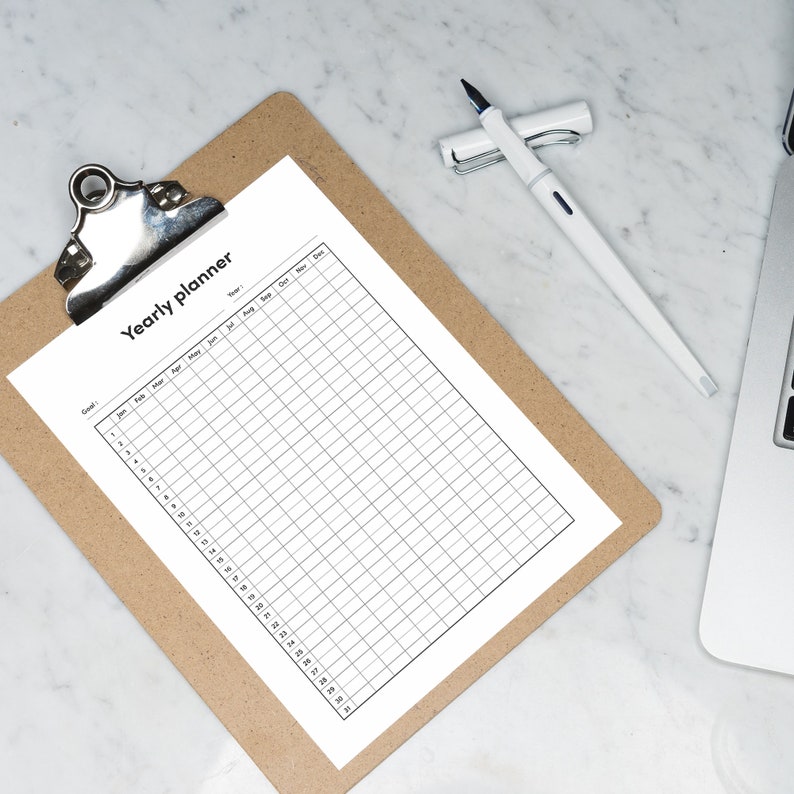 May include: A blank yearly planner on a clipboard with a white pen. The planner has columns for each month of the year and rows for each day of the month.