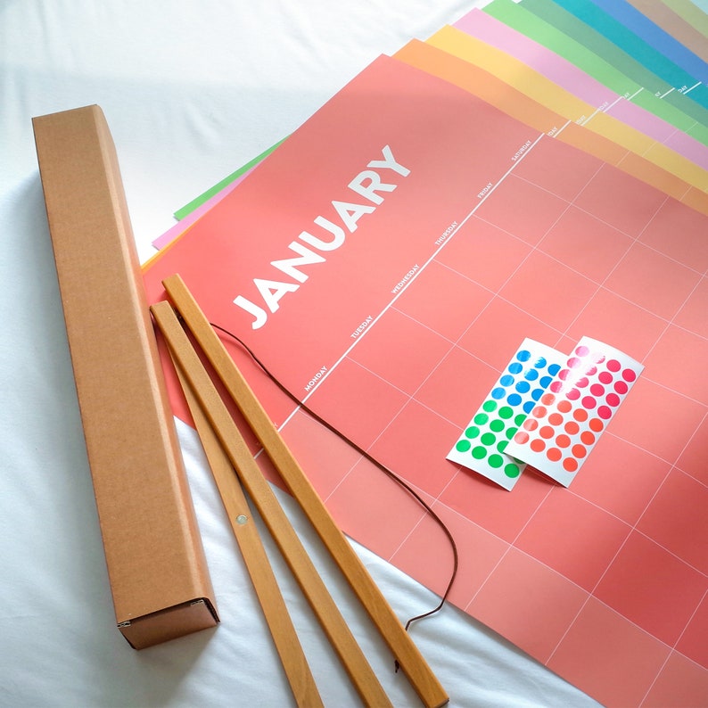 May include: A pink calendar with the word "January" printed in white. The calendar has a grid of squares for each day of the month. There are two sets of colorful dot stickers on the calendar. The calendar is rolled up and has a wooden dowel rod for hanging.