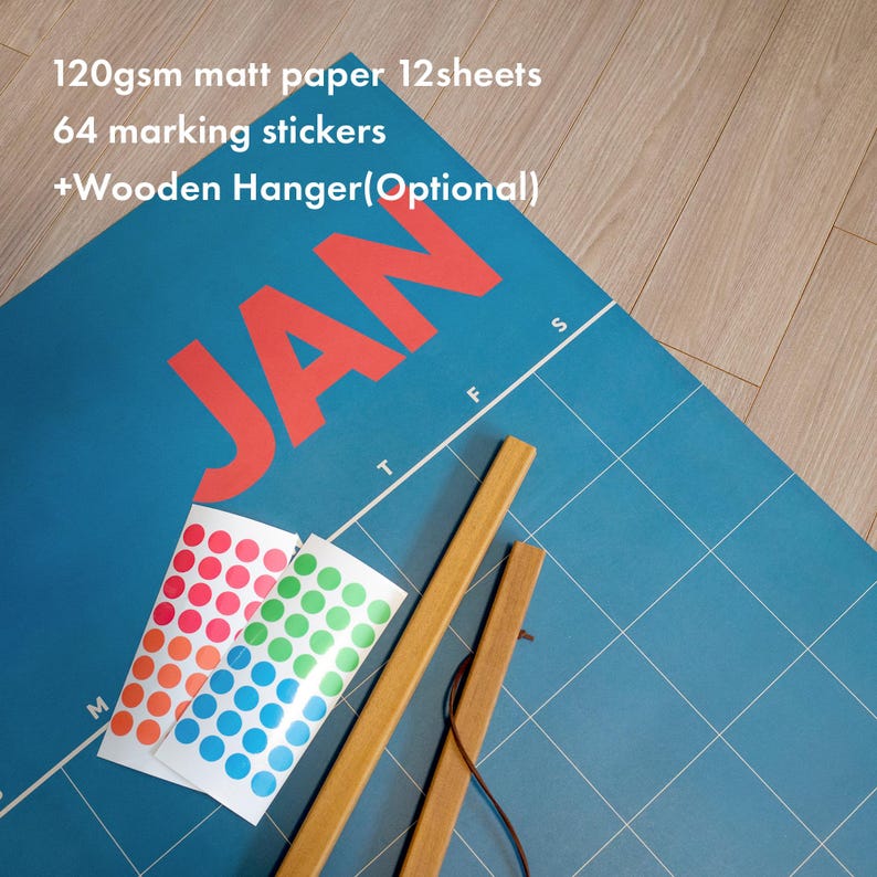 May include: A blue calendar with the month "JAN" in red, featuring a grid pattern and the text "120gsm matt paper 12sheets, 64 marking stickers, +Wooden Hanger(Optional)". Includes colorful dot stickers and wooden hanger.