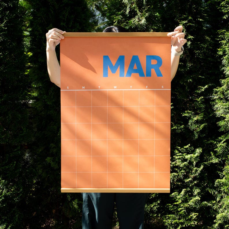 May include: An orange wall calendar for March, with blue text and a grid layout. The calendar is held up by wooden rods at the top and bottom. The background is a lush green foliage.