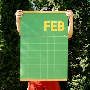 May include: A green wall calendar for February with a grid layout and the word "FEB" in large yellow letters. The calendar is held by a person in front of a green leafy background.