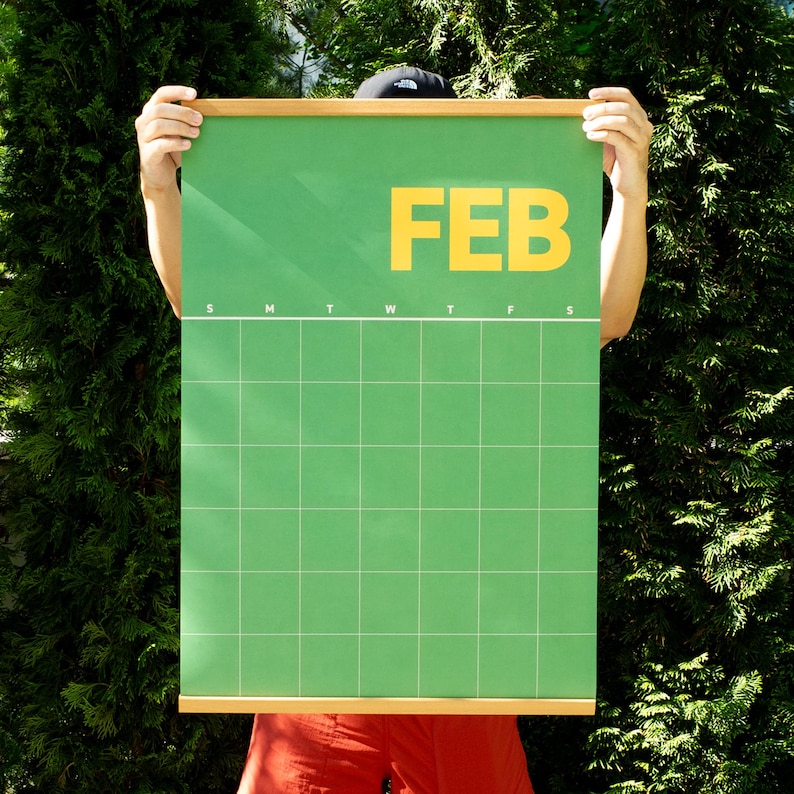 May include: A green wall calendar for February with a grid layout and the word "FEB" in large yellow letters. The calendar is held by a person in front of a green leafy background.
