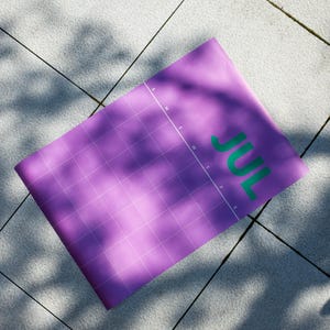 May include: A rolled-up purple calendar with a grid pattern and the month "JUL" in green letters. The calendar is lying on a light grey surface with shadows.