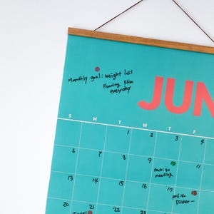 May include: A teal wall calendar for June with a wooden frame and hanging string. The word "JUN" is in large coral letters. Handwritten notes include "Monthly goal: weight loss, Running 5km everyday".