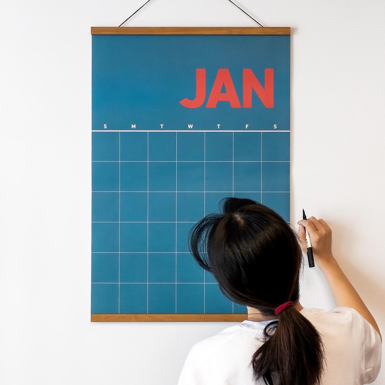 May include: A blue wall calendar displaying "JAN" in red. The calendar grid is visible, with a wooden frame at the top and bottom. A person is writing on the calendar with a black marker.