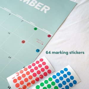 May include: A set of 64 colorful round stickers for marking a calendar. The stickers are in three colors: red, blue, and green. The stickers are shown on a sheet of paper with a calendar printed on it. The calendar has the word "November" at the top and the days of the week listed across the top. The calendar has dates written in black ink. The stickers are arranged in rows and columns on the sheet of paper. The text "64 marking stickers" is written in black ink on the sheet of paper.