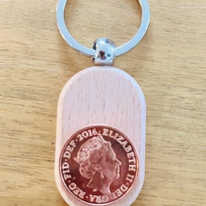 May include: A wooden keychain with a silver-colored ring and a light-colored wooden oval pendant. A copper-colored coin with the profile of a person and the text "ELIZABETH II DEI GRA REG FD 2016" is affixed to the pendant.