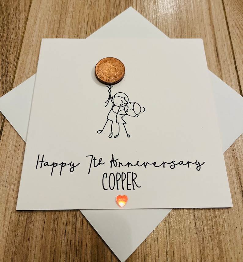 May include: A white greeting card with a copper penny attached, featuring a stick figure couple and the text "Happy 7th Anniversary COPPER." A small, orange heart-shaped gem is at the bottom. The card is inside a white envelope.