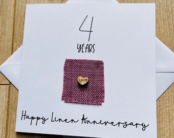 Könnte beinhalten: Eine weiße Karte mit der Zahl 4 und den Worten "Jahre" in schwarzer Tinte. Die Karte trägt auch die Worte "Frohes Leinenjubiläum" in schwarzer Tinte. Ein kleines Holzhäuschen mit dem Wort "Liebe" darauf ist an ein Stück rosafarbenes Jutegewebe befestigt.