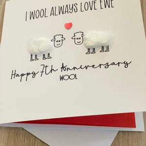 Puede incluir: Tarjeta de felicitaci&oacute;n blanca con el texto "I WOOL ALWAYS LOVE EWE" y "Happy 7th anniversary". La tarjeta presenta dos ovejas hechas de bolas de algod&oacute;n y un peque&ntilde;o coraz&oacute;n rojo. La tarjeta est&aacute; abierta, revelando un interior rojo.