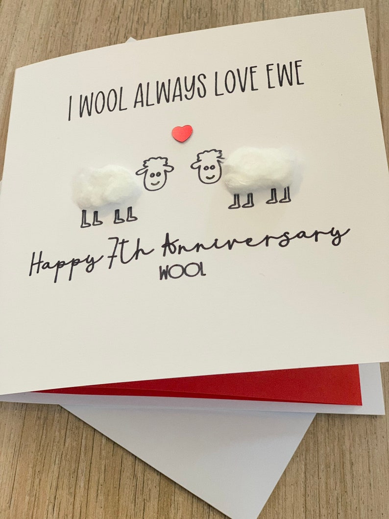 Puede incluir: Tarjeta de felicitaci&oacute;n blanca con el texto "I WOOL ALWAYS LOVE EWE" y "Happy 7th anniversary". La tarjeta presenta dos ovejas hechas de bolas de algod&oacute;n y un peque&ntilde;o coraz&oacute;n rojo. La tarjeta est&aacute; abierta, revelando un interior rojo.