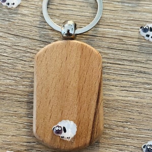 Puede incluir: Un llavero de madera con un anillo plateado y una peque&ntilde;a etiqueta rectangular de madera. La etiqueta presenta un dise&ntilde;o de oveja de dibujos animados. Varios dijes con forma de oveja est&aacute;n esparcidos alrededor del llavero sobre una superficie de madera.