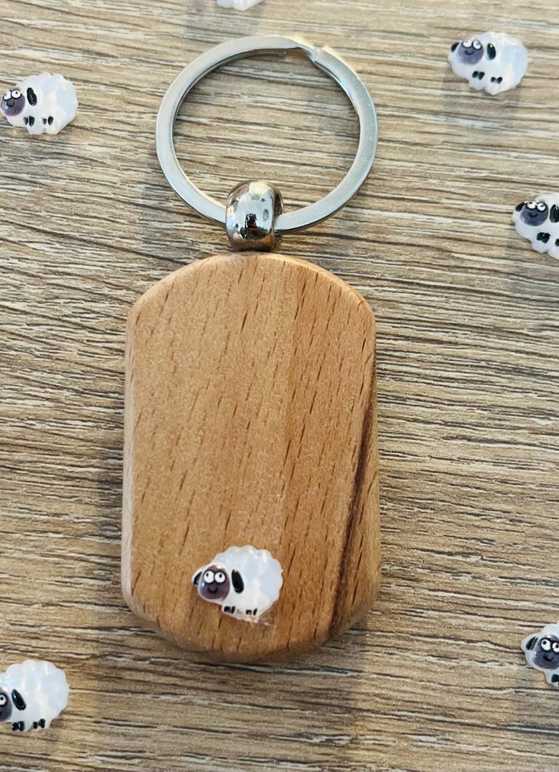 Puede incluir: Un llavero de madera con un anillo plateado y una peque&ntilde;a etiqueta rectangular de madera. La etiqueta presenta un dise&ntilde;o de oveja de dibujos animados. Varios dijes con forma de oveja est&aacute;n esparcidos alrededor del llavero sobre una superficie de madera.