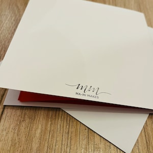 May include: A stack of white greeting cards with a red interior. The cards are on a wooden surface. The bottom card has the text "MA-MI MAKES" in a handwritten style.