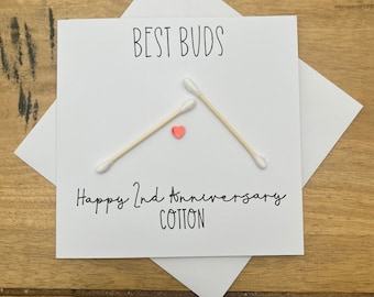Könnte beinhalten: Eine weiße Karte mit schwarzem Text, der "BEST BUDS" und "Happy 2nd Anniversary COTTON" lautet. Drei Wattestäbchen sind in Dreiecksform mit einem kleinen rosa Herzen in der Mitte angeordnet.