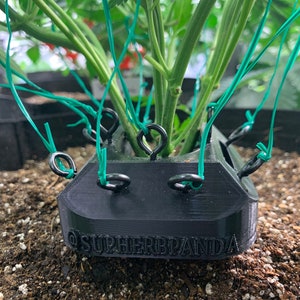 May include: Black plastic plant support with green ties attached to a green plant. The support has the text "@SUPERHERBPANDA" printed on the front.