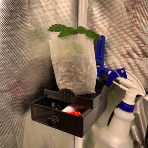 May include: A black 3D printed grow tent accessory with a drawer that says "Super Panda" on it. A small plant in a white grow bag is attached to the accessory with a blue clamp. A white spray bottle with a blue nozzle is also attached to the accessory.