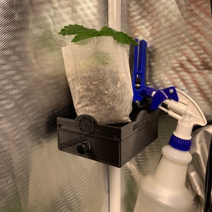 May include: A black plastic plant holder with a small plant in a white fabric pot. The holder has a knob on the front and is attached to a white pole. A clear plastic spray bottle with a blue nozzle is next to the holder.