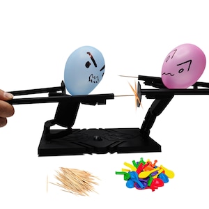 May include: A black plastic toy game with two balloon characters, one blue and one pink, with faces drawn on them. The balloons are attached to a black plastic base with a lever on each side. The balloons are facing each other and have toothpicks sticking out of them. There are also a pile of colorful balloons and a pile of toothpicks on the table.