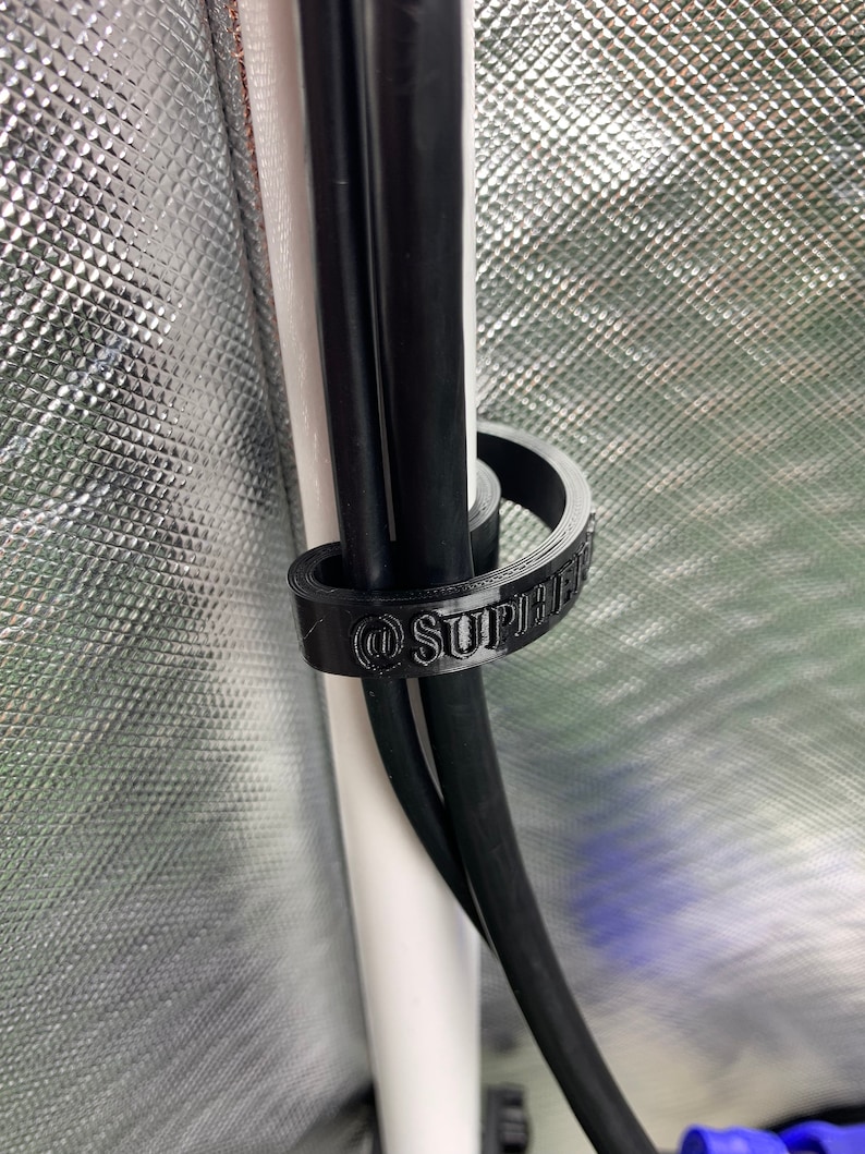 May include: A black cable tie with the text "@SUPREME" printed on it. The cable tie is holding together three black cables.