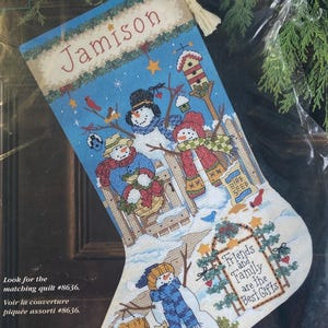 May include: Cross-stitch Christmas stocking with a blue backdrop, depicting snowmen, birds, and a birdhouse. The name "Jamison" is stitched at the top. The phrase "Friends and Family are the Best Gifts" is also stitched on the stocking.