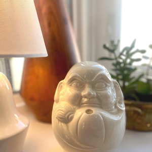 May include: A white ceramic Buddha head figurine with a serene expression. The figurine has a rounded shape and a small hole in the front. Other objects in the background include a lamp, a wooden sculpture, and a small plant.