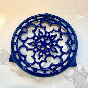 May include: A circular, cobalt blue cast iron trivet featuring an intricate, openwork lattice design with a central star-like motif and three small feet for elevation. The surface has a glossy, smooth finish with symmetrical cutouts throughout.