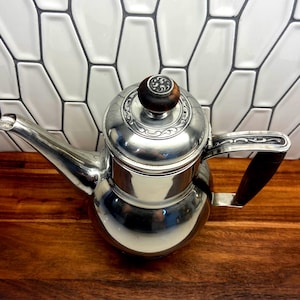 May include: A polished silver teapot with a dark wooden handle and lid knob. The teapot has a curved spout and a decorative design around the lid. The teapot is sitting on a wooden surface.