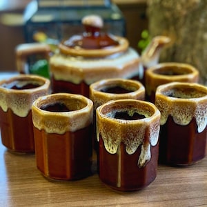 May include: A collection of six brown ceramic mugs with a dripped glaze effect around the rims. A matching teapot with a lid is in the background. The mugs have a hexagonal shape and are arranged on a wooden surface. The set is designed for serving tea or coffee.