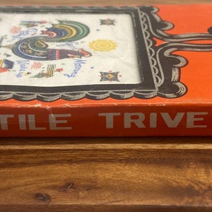 Peut inclure: Un dessous de plat en carreau de fonte encadré dans sa boîte orange d'origine. Le dessous de plat présente un motif de coq coloré sur un carreau blanc. La boîte est étiquetée "TILE TRIVET FRAMED".