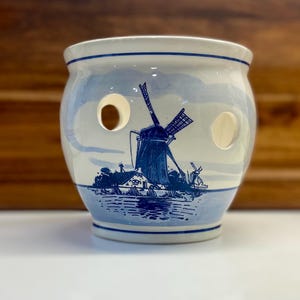May include: A round, white and blue ceramic onion keeper with three circular holes. The design features a blue windmill scene with a water landscape. The keeper has a blue rim and base.