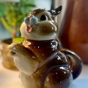 Peut inclure: Une figurine en céramique brillante représentant un écureuil brun et blanc avec une patte levée, une petite langue rose visible et des yeux peints foncés. L'écureuil a un corps arrondi, une queue touffue et une finition lisse et réfléchissante.