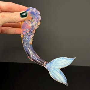 May include: A glass mermaid tail pipe with a pink and blue iridescent finish. The tail is detailed with a textured, bubble-like design and a white, iridescent fin.