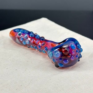 May include: A glass pipe with a red and blue swirl pattern and a textured surface. The pipe is covered in small, raised bubbles.