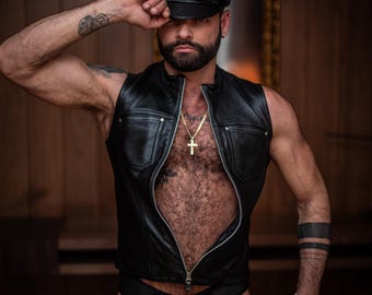 May include: Black leather vest with a zipper, paired with a matching cap and briefs. A gold cross necklace adds a touch of detail. The outfit is completed with tattoos on the arms and leg.