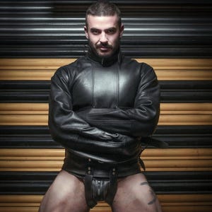 May include: A person wearing a black leather jacket with the sleeves rolled up and a black leather harness. The person is standing in front of a black and yellow striped garage door with a sign that reads "MAXIMUM VEHICLE HEIGHT".