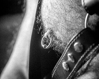 May include: A close-up of a person's torso with a silver ring piercing in their chest. The person is wearing a leather harness with metal accents.