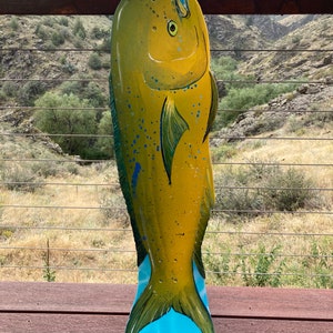 May include: A yellow and blue painted wooden fish sculpture of a mahi-mahi, also known as a dorado, with a blue tail fin. The sculpture is standing upright on a wooden deck.