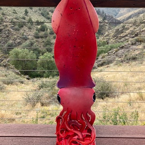 May include: A red and purple painted wooden squid sculpture with a long body and tentacles. The sculpture is standing on a wooden deck.
