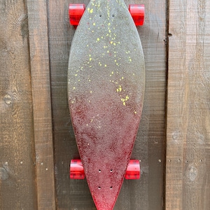 May include: A red, yellow, and black longboard skateboard with red wheels. The skateboard is hanging on a wooden wall.
