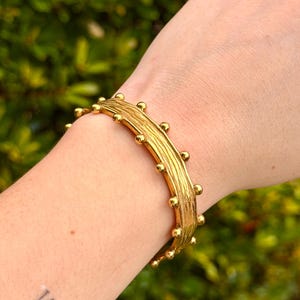 May include: A gold-toned cuff bracelet featuring a textured, bark-like surface finish. The edges of the bracelet are adorned with a series of small, evenly spaced spherical studs that run along both the top and bottom borders of the piece.