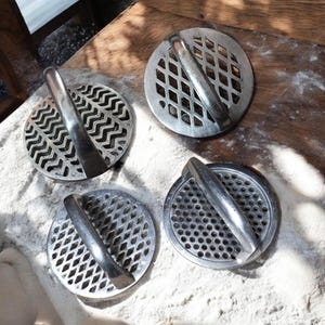 May include: Four silver-colored, circular kitchen tools with handles. Each tool has a different patterned design on the circular base, including zigzags, diamond shapes, and small holes. The tools are arranged on a wooden surface dusted with a white powder.