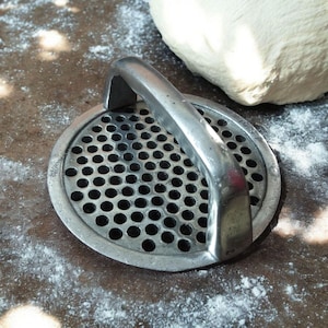 May include: A metal spaetzle maker with a perforated base and a curved handle. The tool is silver and sits on a surface dusted with flour, with a ball of dough in the background. This kitchen utensil is used for making spaetzle.