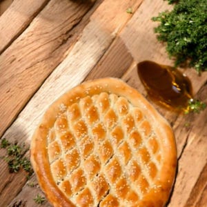 May include: A round, golden-brown flatbread with a diamond pattern and sesame seeds, presented on a wooden surface. The image also includes a small amount of herbs and a pool of oil.