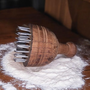 Puede incluir: Un ablandador de carne de madera con púas metálicas, apoyado sobre una superficie espolvoreada con harina. El ablandador tiene una cabeza redondeada y un mango, mostrando la veta natural de la madera.