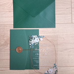 Peut inclure: Une invitation de mariage en acrylique transparente avec un cadre géométrique doré et un motif floral. Le texte de l'invitation indique "Anastasia & Romain" et comprend la date "Samedi 25 mai 2024".