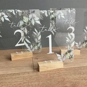 Peut inclure: Trois numéros de table en acrylique transparent avec un motif floral aquarelle dans des tons de vert et de blanc. Les chiffres sont blancs et le texte "table" est écrit en blanc en cursive. Chaque signe est sur un support en bois.