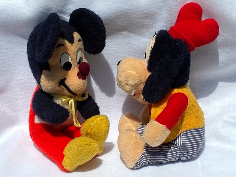 May include: Two vintage plush toys, a black and white mouse wearing red pants and a yellow bow, and a black and white dog wearing a red hat, yellow shirt, and blue and white striped pants.