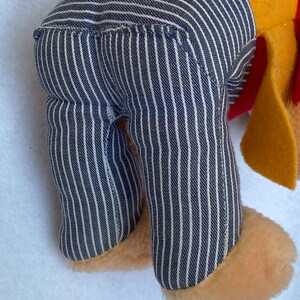 May include: A plush toy with a yellow body, blue and white striped pants, and a red collar. The toy is standing on a white surface.